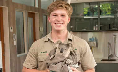 Young man smiling while holding a koala wrapped in a blanket indoors.