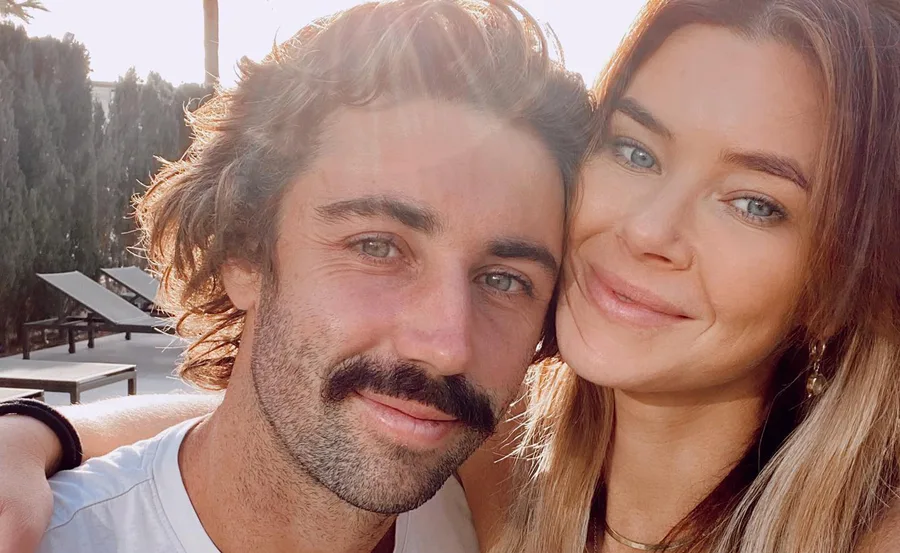 A couple taking a close-up selfie outdoors, smiling with sunlight in the background.