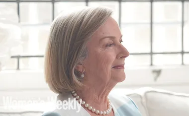 An elderly woman smiles while sitting indoors, wearing pearl earrings and necklace. "The Australian Women's Weekly" text overlay.