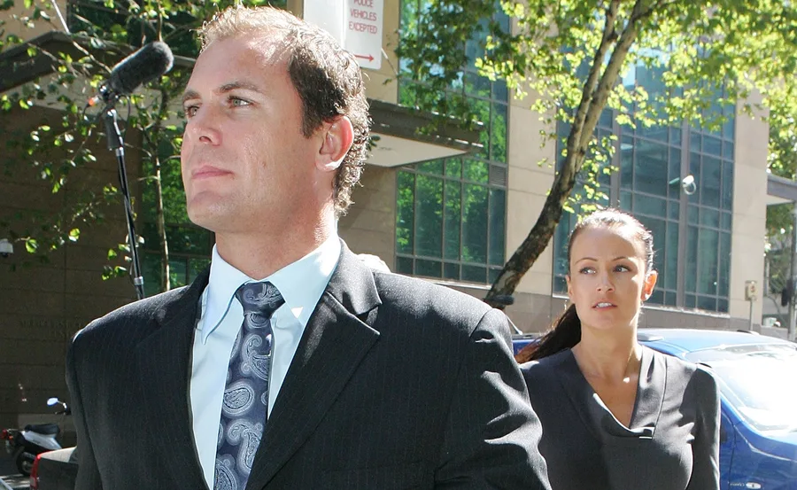 A man in a suit walks in front of a woman on a city street, with a microphone visible above them.