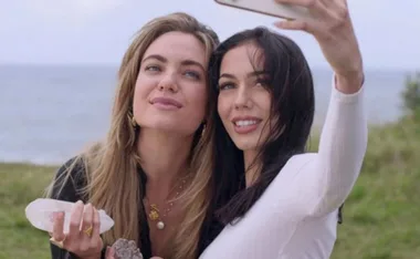 Two women taking a selfie by the sea, one holding crystals, both smiling and looking content.