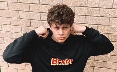 Young man with curly hair wearing a black "Brxtn" hoodie, grasping the hood, stands in front of a brick wall.