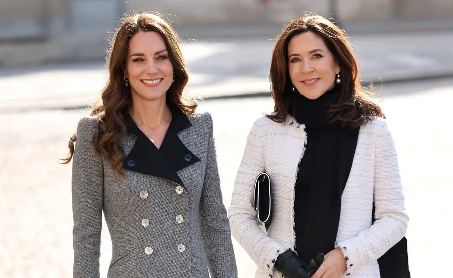 Two women smiling outdoors, one in a gray coat and the other in a white jacket, holding a clutch.