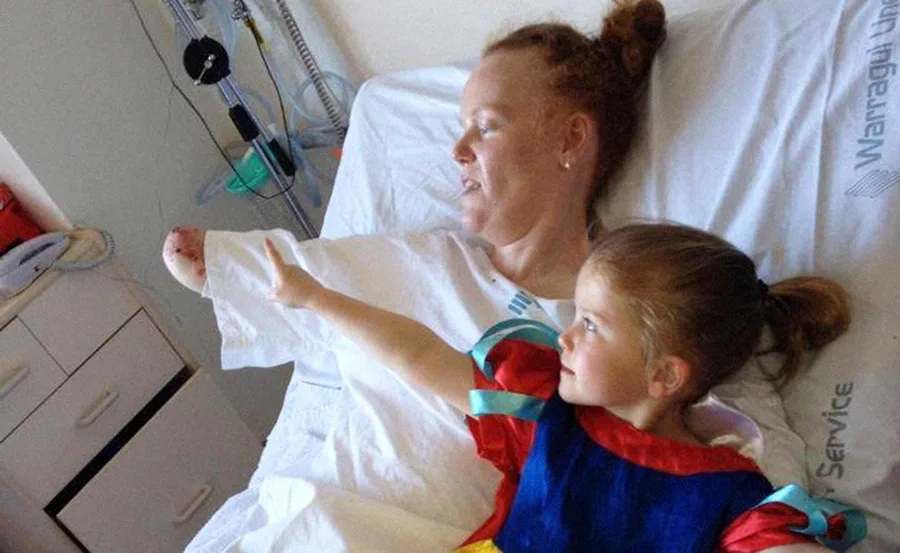 Mother with amputated arm lies in hospital bed, daughter in Snow White costume beside her, both reaching out and smiling.