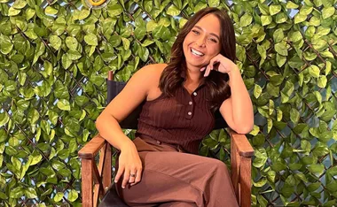 Woman in brown outfit sitting on a director's chair, smiling against a backdrop of green leaves.