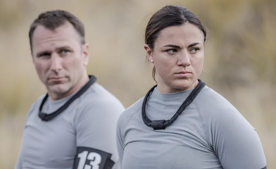 Two people in gray shirts outdoors, both wearing armbands; one appears focused while the other looks to the side.