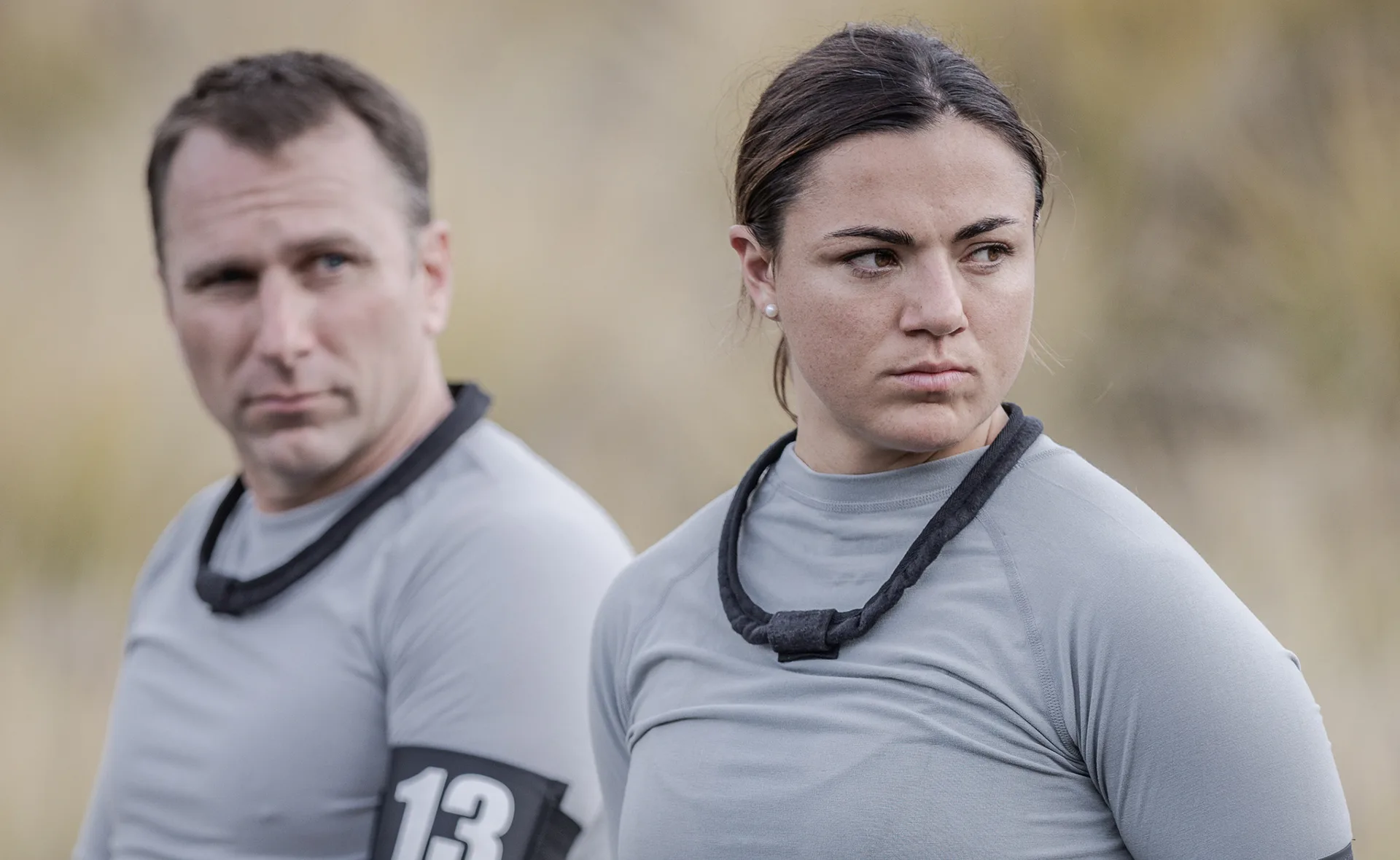 Two people in gray shirts outdoors, both wearing armbands; one appears focused while the other looks to the side.