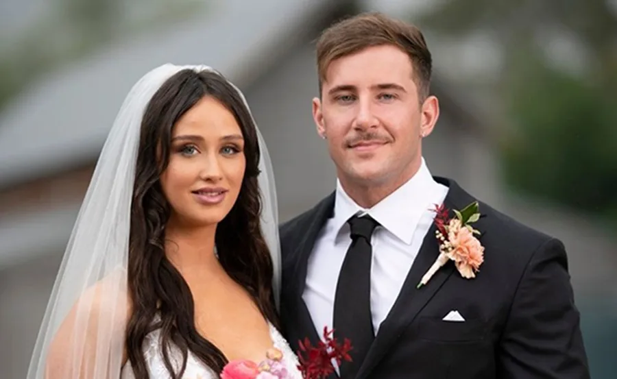 Bride and groom pose outdoors, bride in white dress and veil, groom in black suit with boutonniere.
