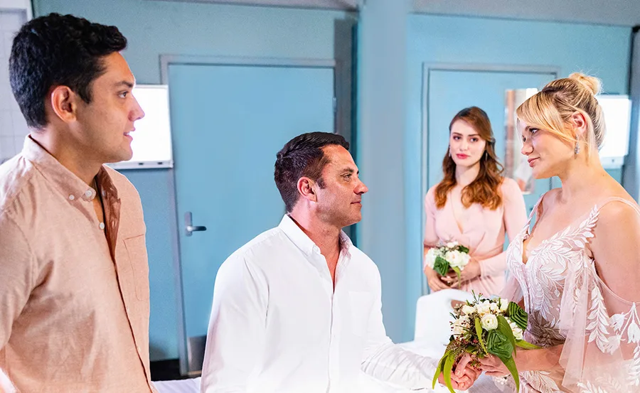 Mia in a bridal gown holds a bouquet, standing with Ari and others in an emotional scene from "Home and Away."