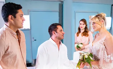 Mia in a bridal gown holds a bouquet, standing with Ari and others in an emotional scene from "Home and Away."