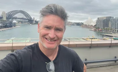 A person taking a selfie with the Sydney Harbour Bridge and Opera House in the background on a cloudy day.