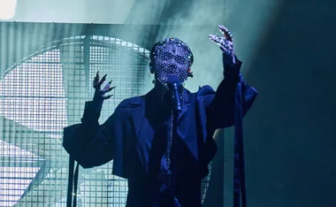 Sheldon Riley performs in a dark outfit and ornate face mask, singing into a microphone under dramatic lighting.