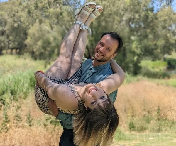 Couple posing outdoors; man holds woman upside down playfully, both smiling, with a sunny, leafy background.