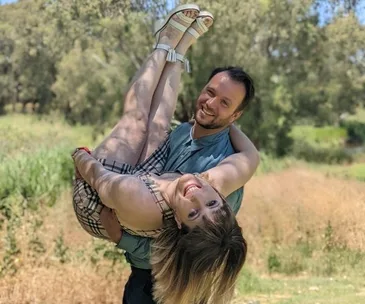 Couple posing outdoors; man holds woman upside down playfully, both smiling, with a sunny, leafy background.