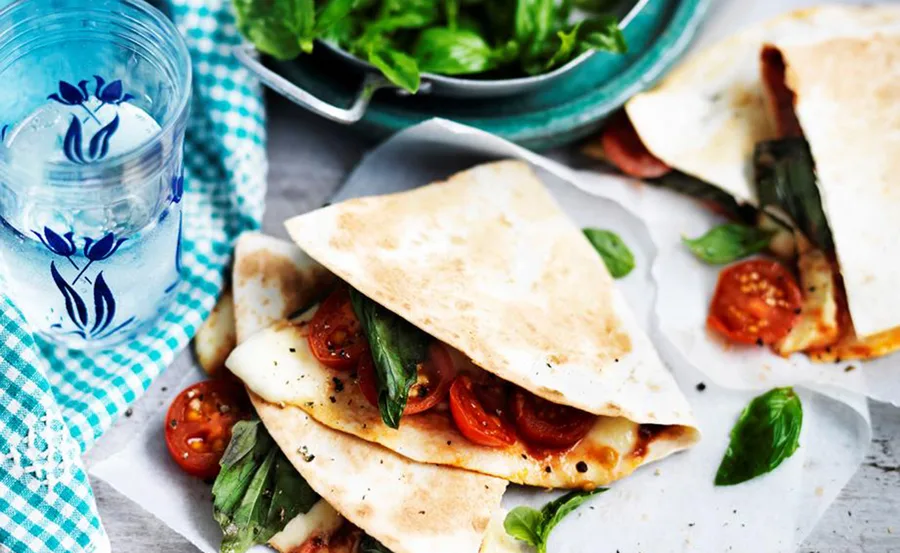 Quesadillas filled with cheese, tomatoes, and basil on a plate, next to a glass of water and fresh basil leaves.
