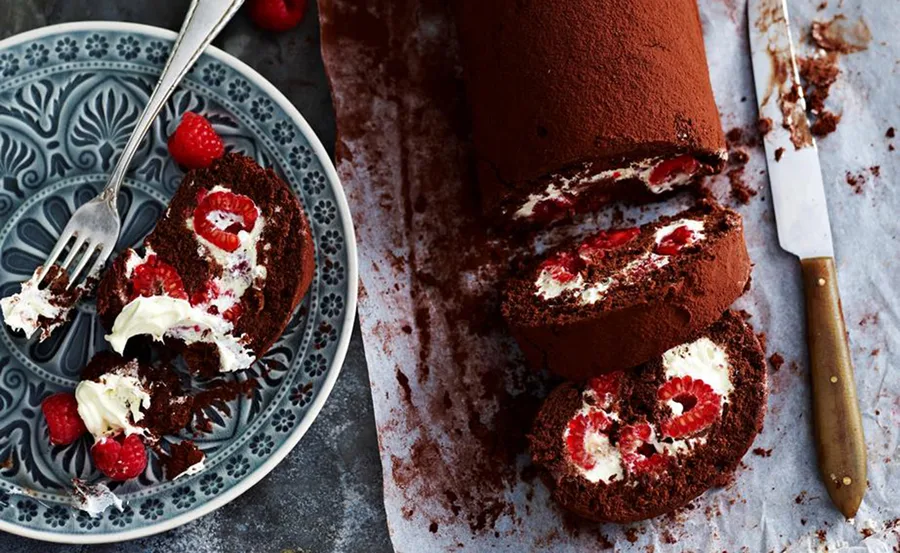 Chocolate roulade with whipped cream and raspberries, partially sliced, displayed on a patterned plate with a fork.