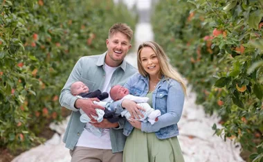 A couple smiling and holding two babies in a lush orchard path.