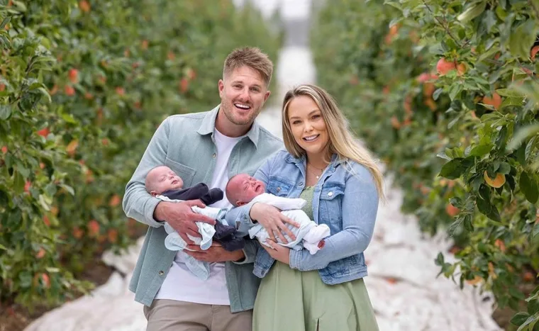 A couple smiling and holding two babies in a lush orchard path.