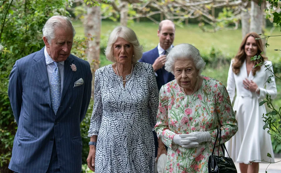 A group of elegantly dressed individuals walk in a garden setting, engaged in conversation.