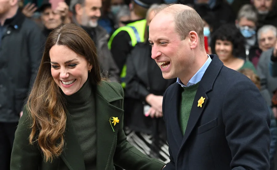 Prince William and Kate Middleton smiling, wearing daffodils, surrounded by people.