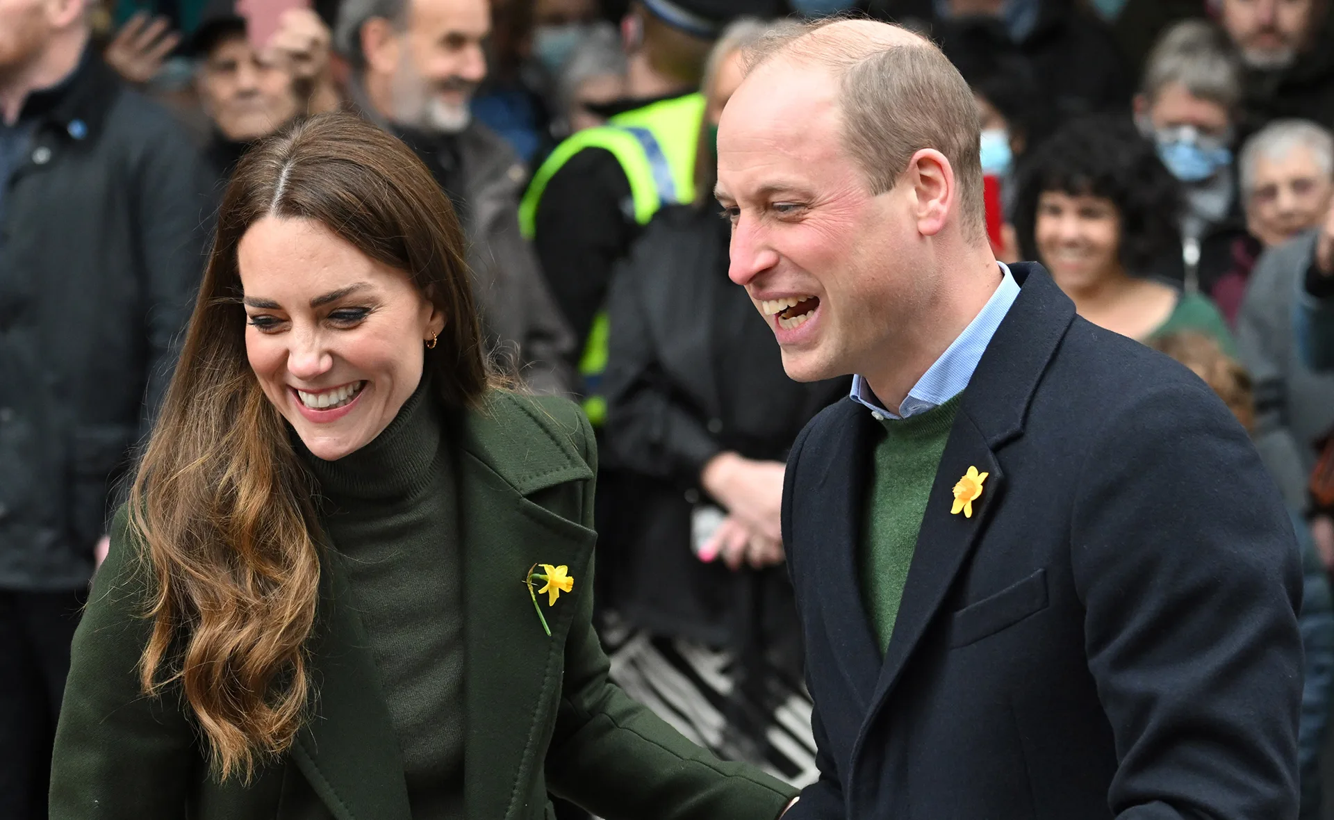 Prince William and Kate Middleton smiling, wearing daffodils, surrounded by people.