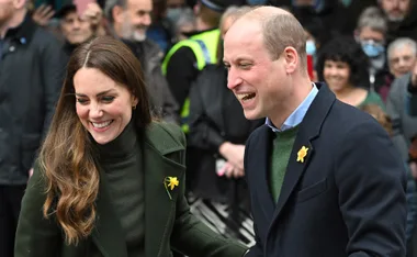 Prince William and Kate Middleton smiling, wearing daffodils, surrounded by people.