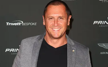 A smiling man in a black shirt and gray blazer at a formal event with dark background and various logos.