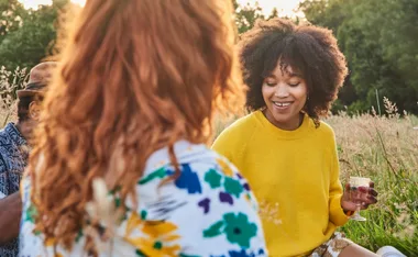 Two people enjoying a picnic outdoors, one wearing a yellow sweater and holding a glass, smiling in a sunny field.