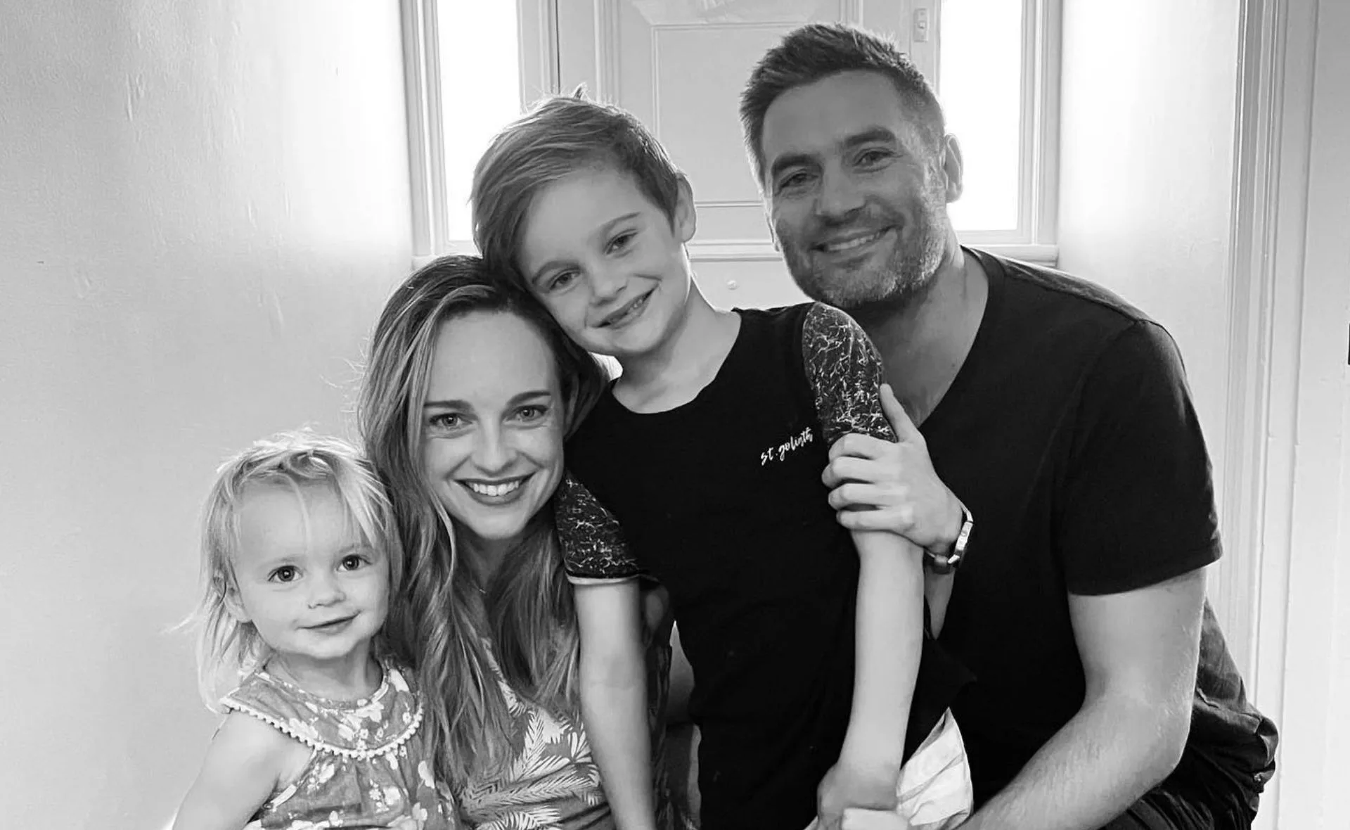 A smiling family of four poses closely together in a well-lit room, showcasing a warm, happy moment.