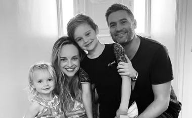 A smiling family of four poses closely together in a well-lit room, showcasing a warm, happy moment.