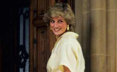 Princess Diana smiling, wearing a yellow blouse, standing outside a building with wooden doors.