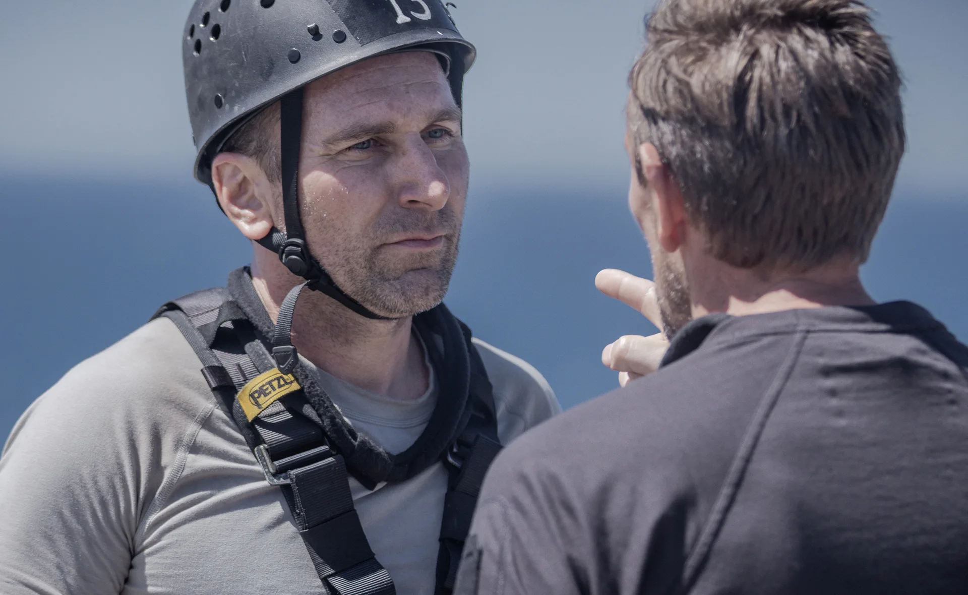 A person wearing a helmet and harness listens intently to someone pointing, against a blue sky backdrop.