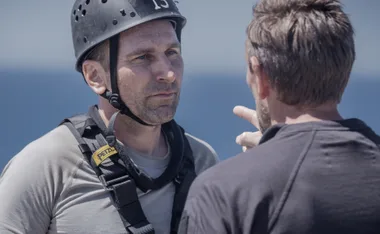 A person wearing a helmet and harness listens intently to someone pointing, against a blue sky backdrop.