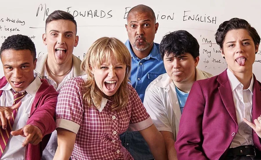 Group of teens and a teacher in a classroom, pulling funny faces and posing energetically in front of a whiteboard.