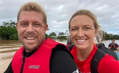 Two people in red life vests smiling during a boat ride on a flooded area, with trees and cloudy sky in the background.