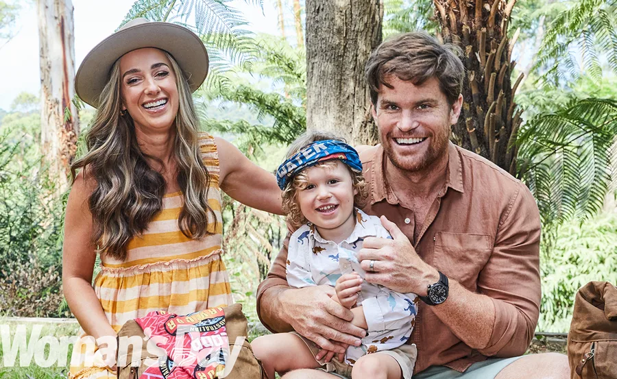 Family of three smiling outdoors, man holding child on lap, woman wearing striped dress and hat, surrounded by trees.