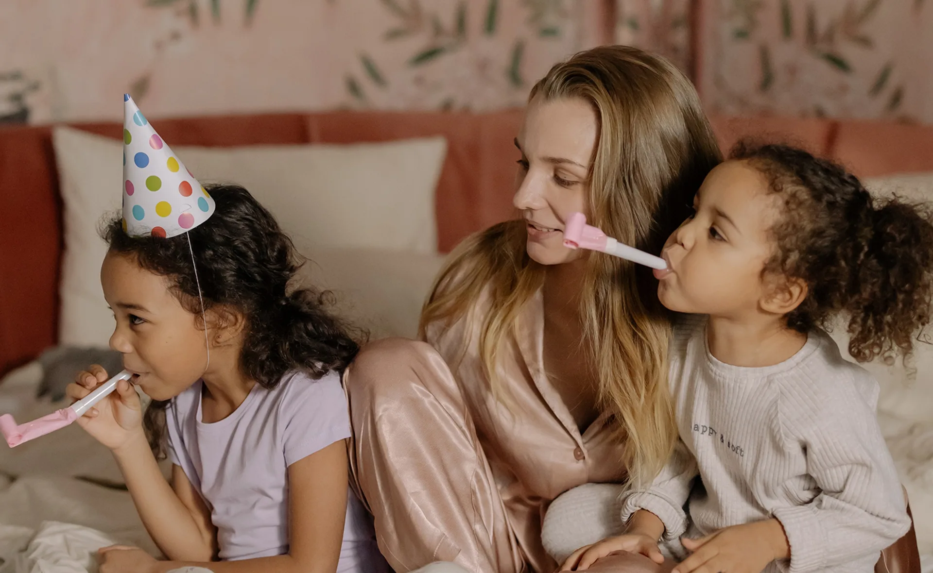 A woman and two children on a bed celebrate with party hats and noisemakers.
