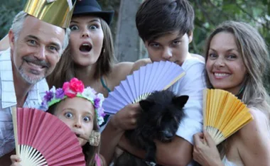 A happy family posing with colorful fans and wearing fun hats, smiling and looking playful outdoors.