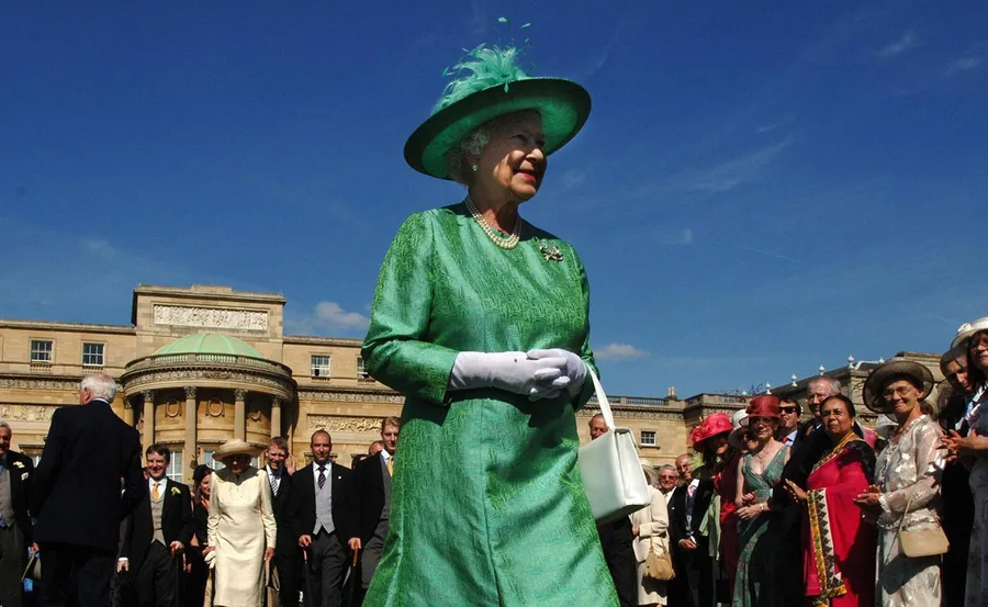 A woman in a green outfit and hat at a formal outdoor event with people gathered in front of a large building.