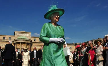 A woman in a green outfit and hat at a formal outdoor event with people gathered in front of a large building.