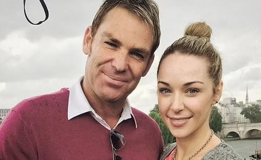 A smiling man in a red sweater with sunglasses and a woman with a bun, posing outdoors on a cloudy day.