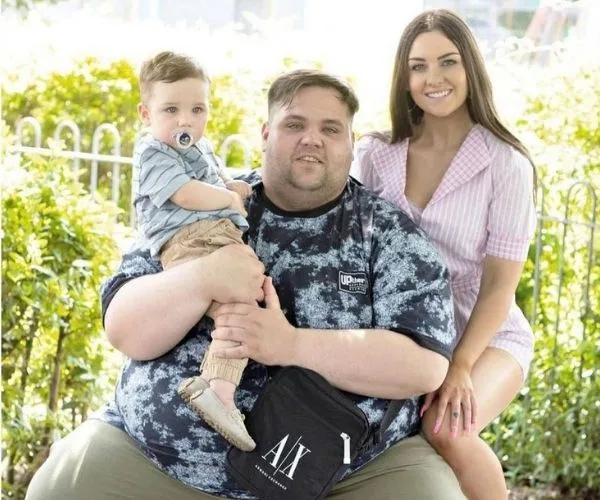 Family posing in a park; man seated holding a baby with a pacifier, woman behind smiling, greenery in the background.
