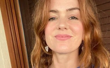 A person with reddish-brown hair smiles softly, wearing heart-shaped earrings in natural light.