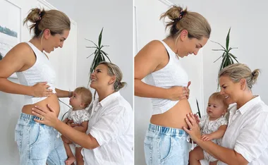 Two women smiling, one pregnant, with a baby on the other's lap, indoors with a plant in the background.