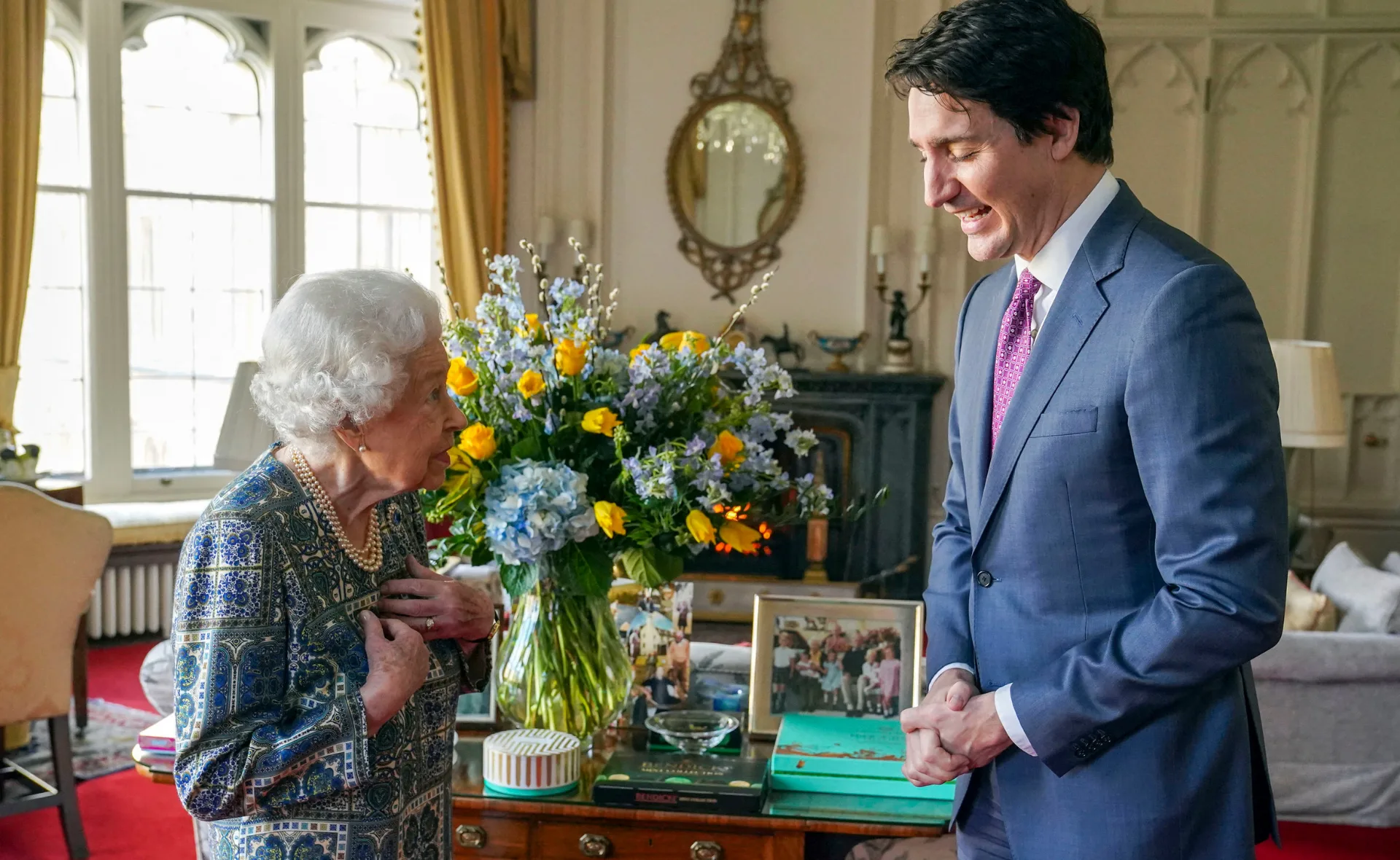 Two individuals smiling and conversing in an elegantly decorated room with flowers and framed photos on a table.