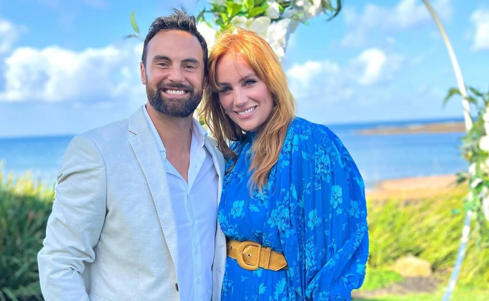 Couple posing together, smiling outdoors near the beach, with blue sky and greenery in the background.