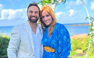 Couple posing together, smiling outdoors near the beach, with blue sky and greenery in the background.