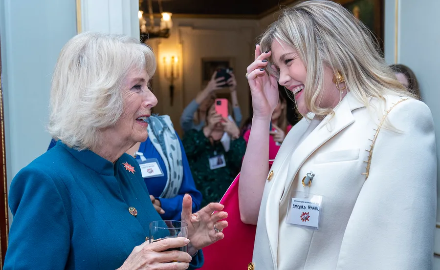 Two women smiling and talking at an event, one holding a drink. Others in the background are taking photos.
