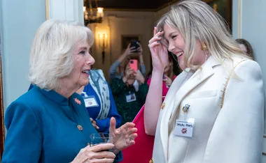 Two women smiling and talking at an event, one holding a drink. Others in the background are taking photos.