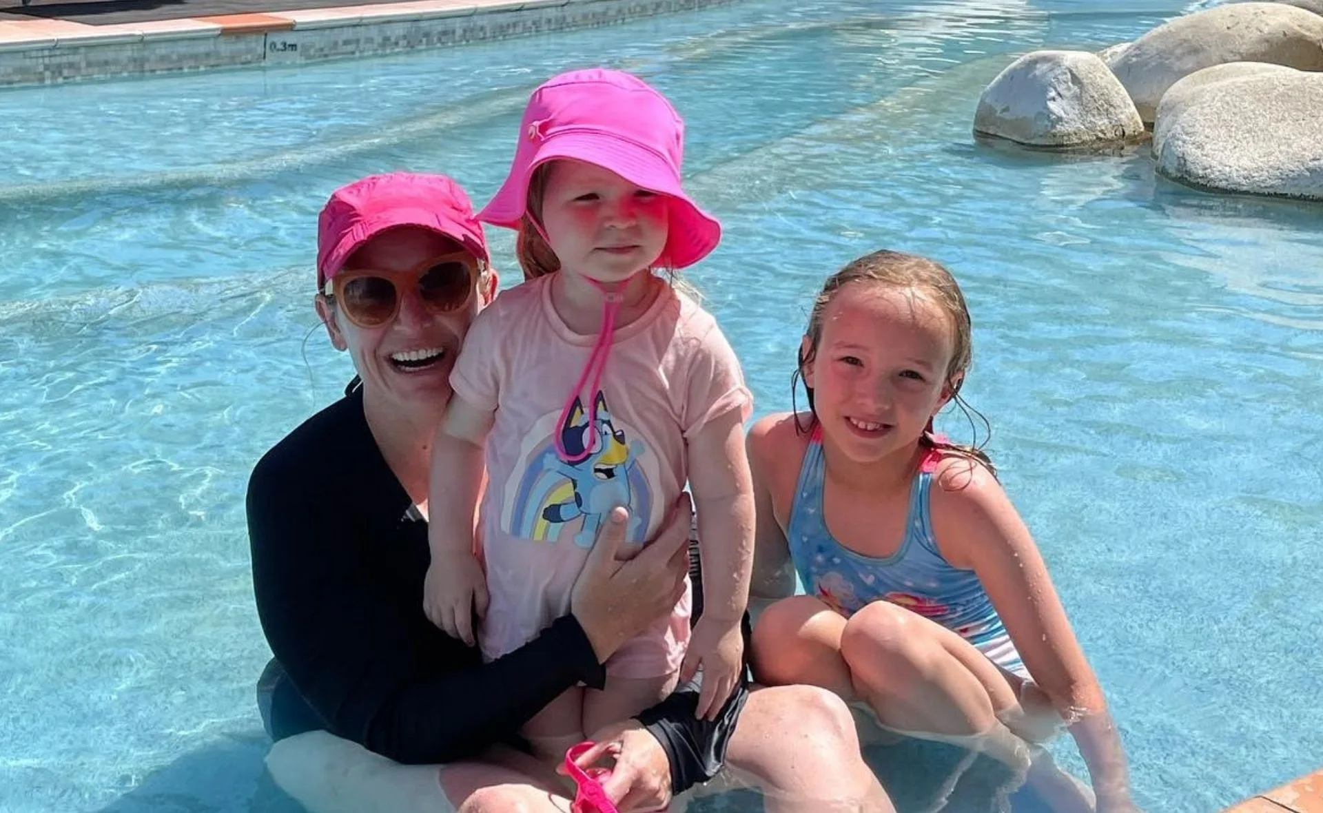 A woman and two children in a pool, wearing pink hats and swimsuits, smiling on a sunny day.